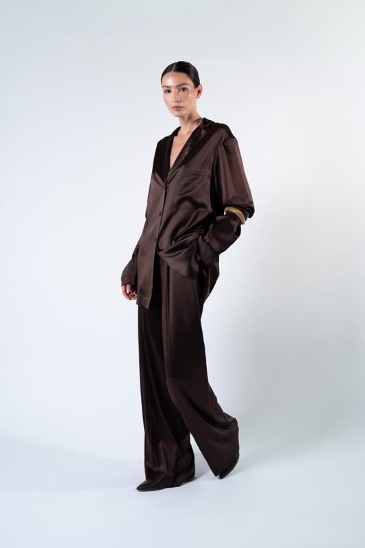 Modella fotografata a tre quarti su sfondo chiaro mentre indossa una camicia oversize in satin di seta marrone con rever, taschino applicato sul petto, chiusura a tre bottoni e maniche lunghe. La camicia è abbinata a pantaloni coordinati in satin di seta, con una mano nella tasca del pantalone per evidenziare la linea morbida e la vestibilità rilassata del completo.