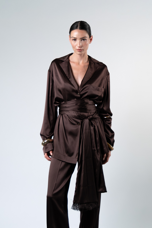 Modella fotografata frontalmente su sfondo chiaro mentre indossa una camicia oversize in satin di seta marrone con rever, taschino applicato e chiusura a tre bottoni, abbinata a pantaloni coordinati. La camicia è parzialmente annodata in vita con una fusciacca dello stesso tessuto, creando un effetto drappeggiato sul davanti.