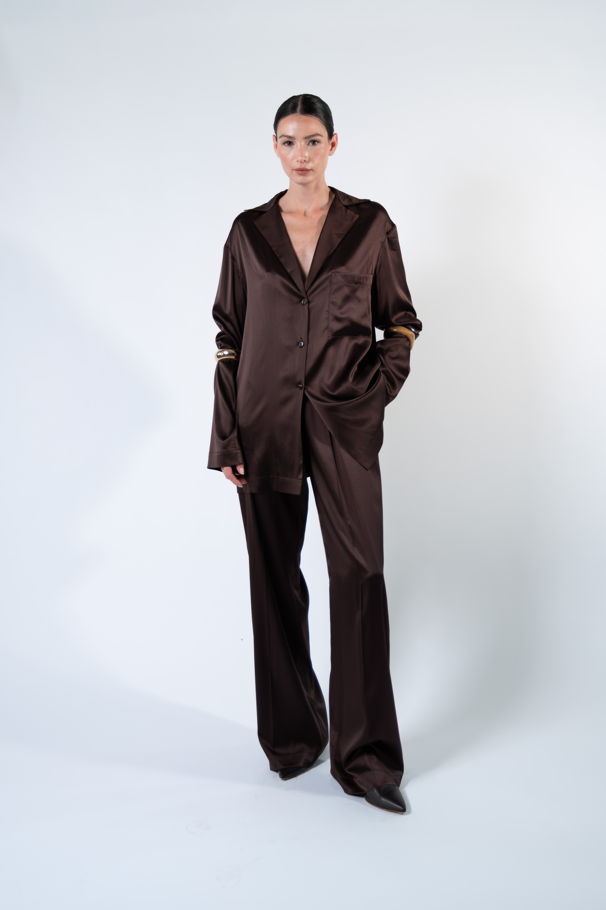 Modella fotografata frontalmente su sfondo chiaro mentre indossa una camicia oversize in satin di seta marrone con rever, taschino applicato sul petto, chiusura a tre bottoni e maniche lunghe. La camicia è abbinata a pantaloni coordinati in satin di seta, con una mano nella tasca del pantalone per evidenziare la linea morbida e la vestibilità rilassata del completo.