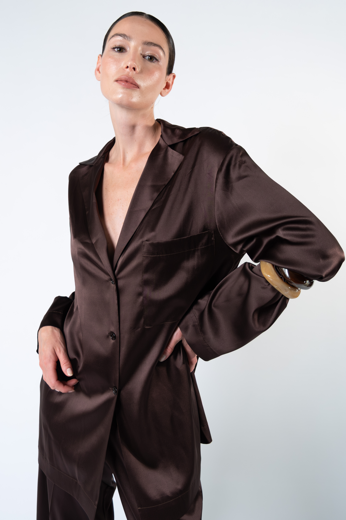 Primo piano di una modella che indossa una camicia oversize in satin di seta marrone con rever, taschino applicato sul petto, chiusura frontale a tre bottoni e maniche lunghe. L'inquadratura mette in risalto la lucentezza naturale del satin di seta, la fluidità del tessuto e la costruzione morbida della silhouette