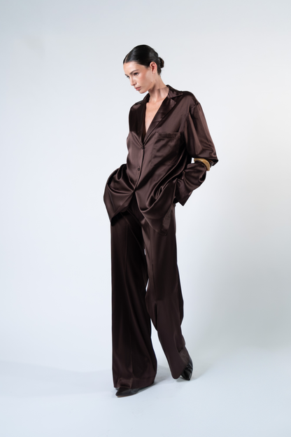 Modella fotografata a tre quarti su sfondo chiaro mentre indossa una camicia oversize in satin di seta marrone con rever, taschino applicato sul petto, chiusura a tre bottoni e maniche lunghe. La camicia è abbinata a pantaloni coordinati in satin di seta, con una mano nella tasca del pantalone per evidenziare la linea morbida e la vestibilità rilassata del completo.