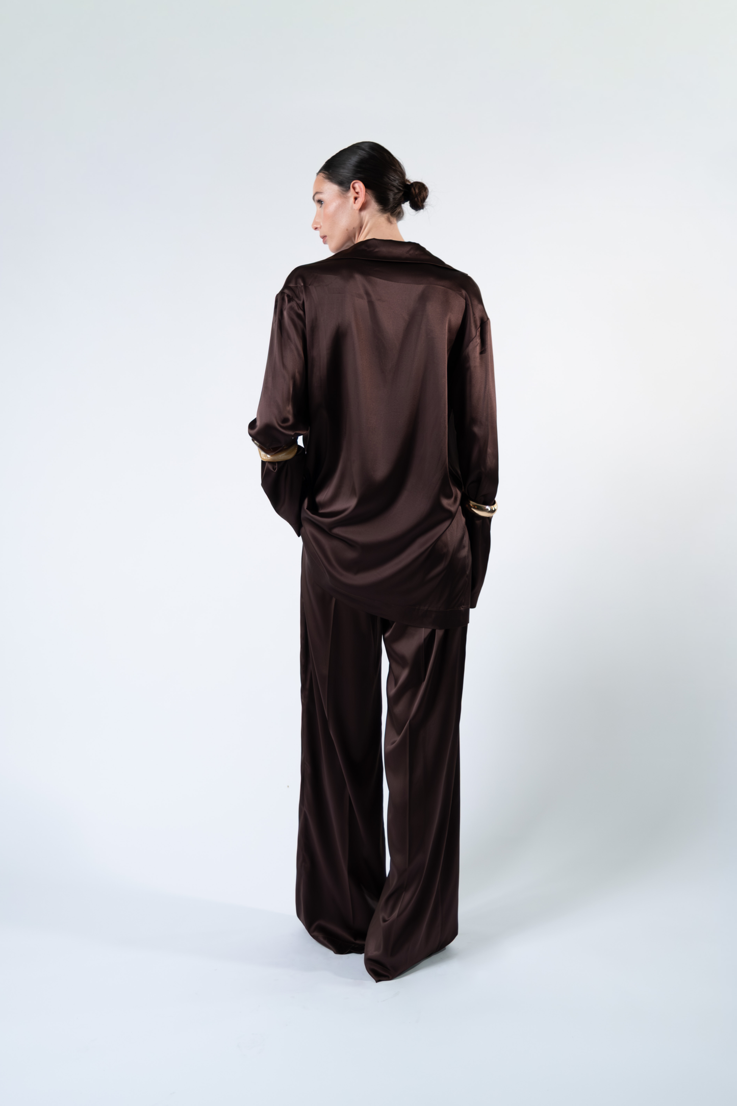 Modella fotografata dal retro su sfondo chiaro mentre indossa una camicia oversize in satin di seta marrone a maniche lunghe. La camicia è abbinata a pantaloni coordinati in satin di seta, con una mano nella tasca del pantalone per evidenziare la linea morbida e la vestibilità rilassata del completo.