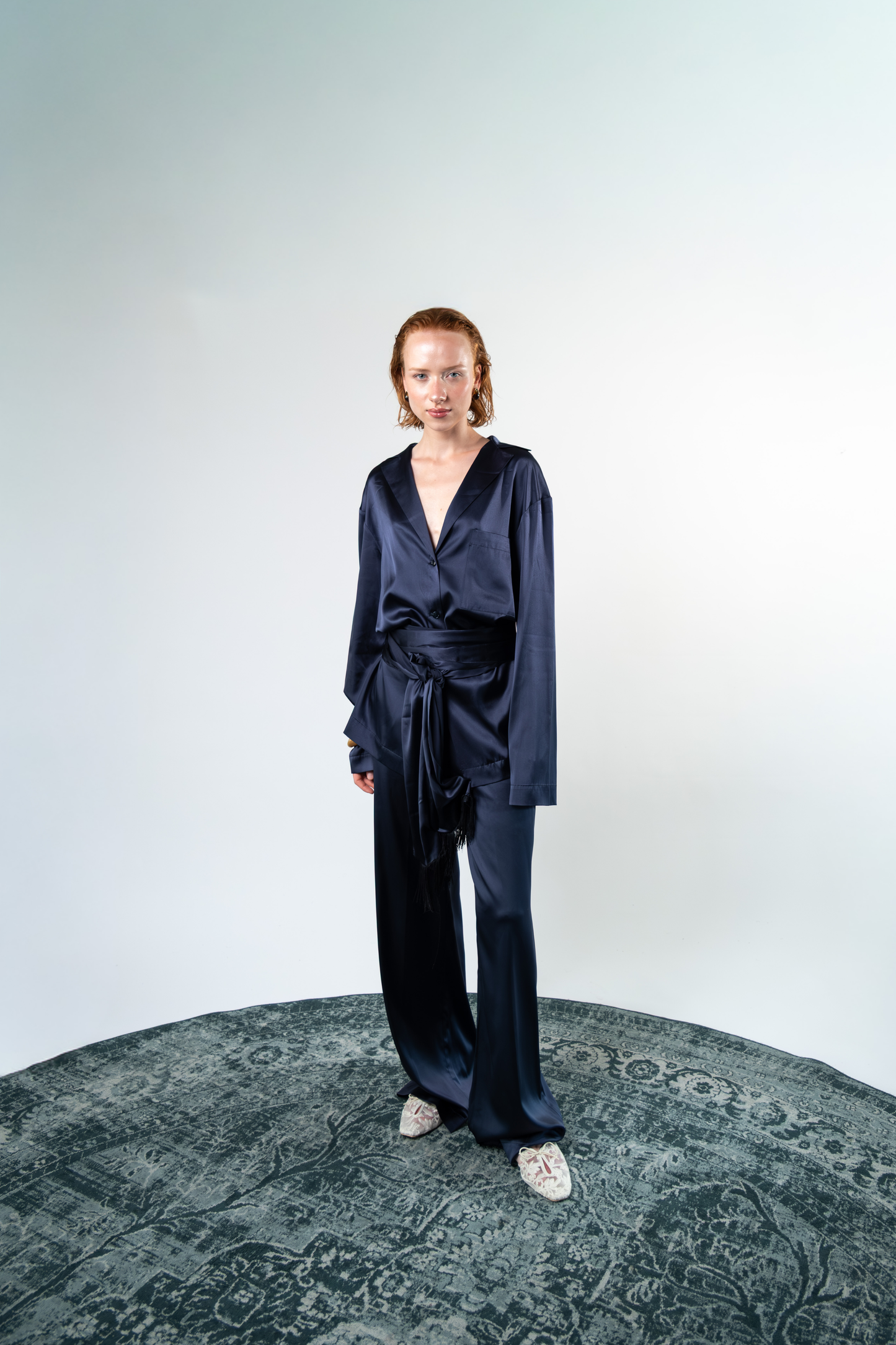 Camicia oversize in satin di seta blu abbinata al pantalone coordinato e completata da una fascia annodata in vita. La silhouette morbida valorizza la fluidità del tessuto e l'ispirazione tailoring del rever.