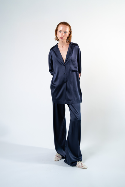 Vista frontale della camicia oversize in satin di seta blu con rever, taschino applicato e chiusura con tre bottoni. La silhouette lunga e morbida valorizza la lucentezza e il drappeggio naturale del satin.