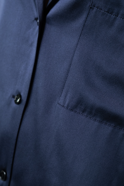 Dettaglio del taschino applicato e della chiusura con bottoni della camicia oversize in satin di seta blu. La fotografia mette in risalto la superficie liscia del satin e la precisione delle cuciture.